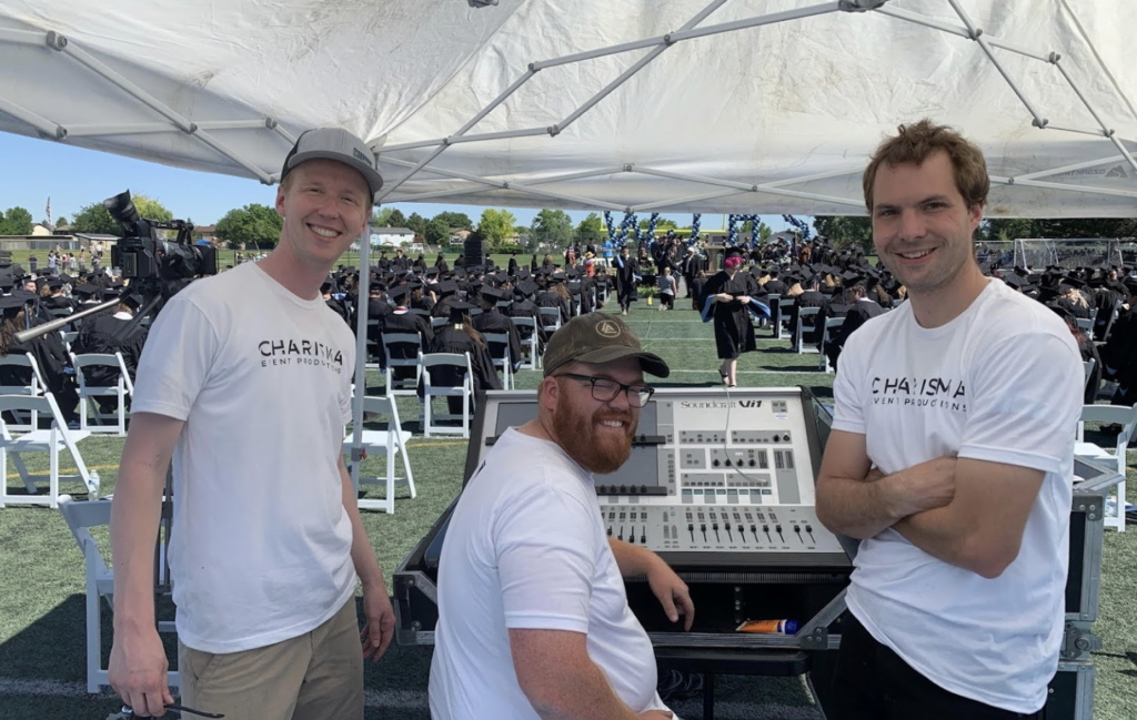 Three male sound engineers smiling in white t-shirts while working an outside graduation ceremony.