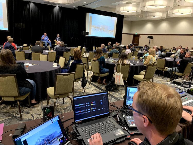 An engineer sitting at a laptop while two men speak on stage to a room of people.