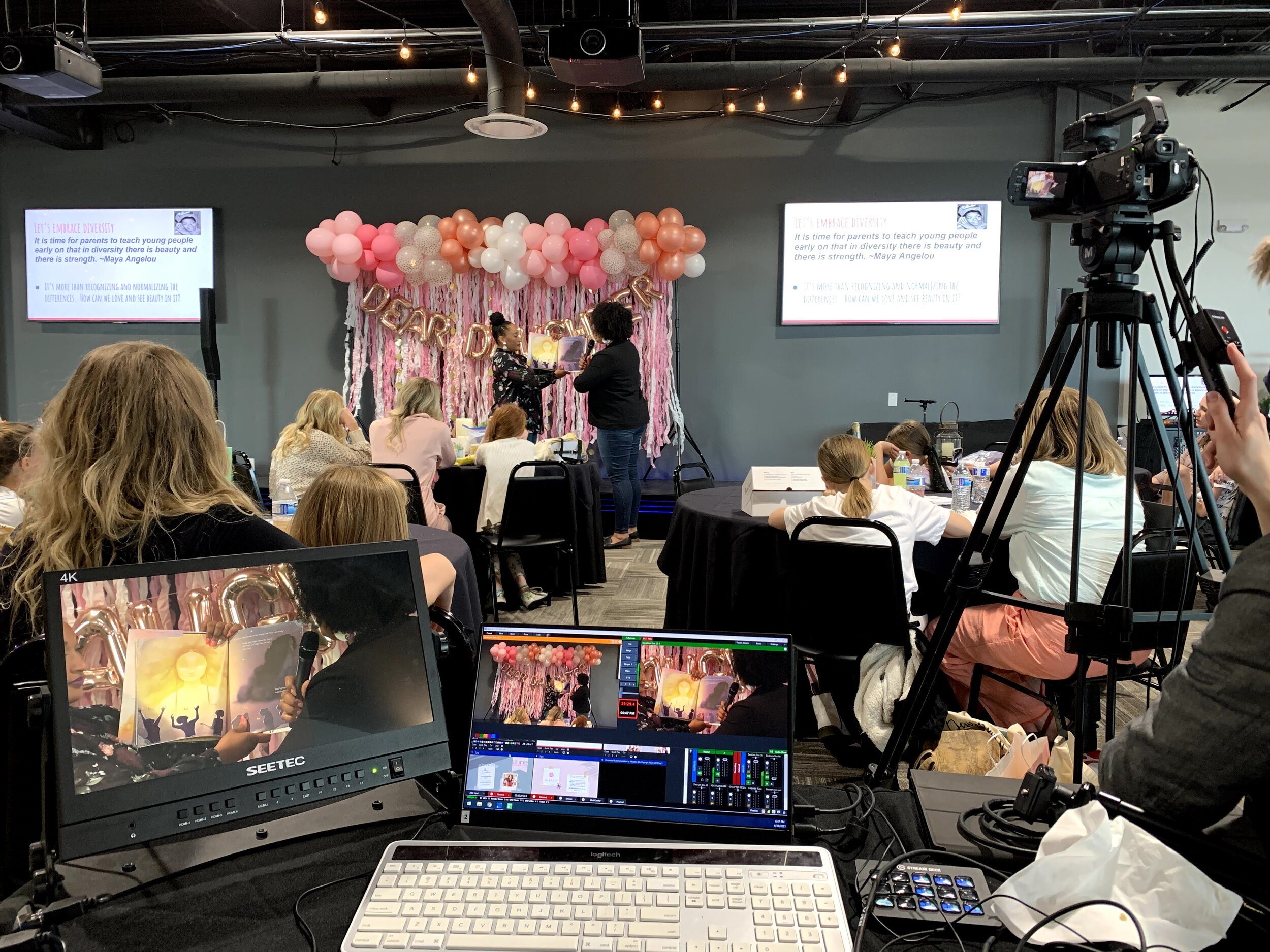 Two women showcase a picture book in front of an audience and camera.