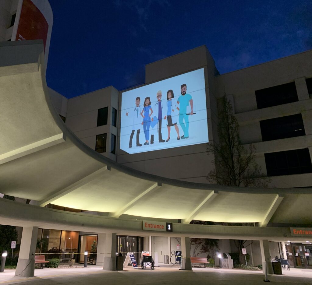 The outdoor entrance area of a hospital with a projected image of animated medical workers up higher on the building.