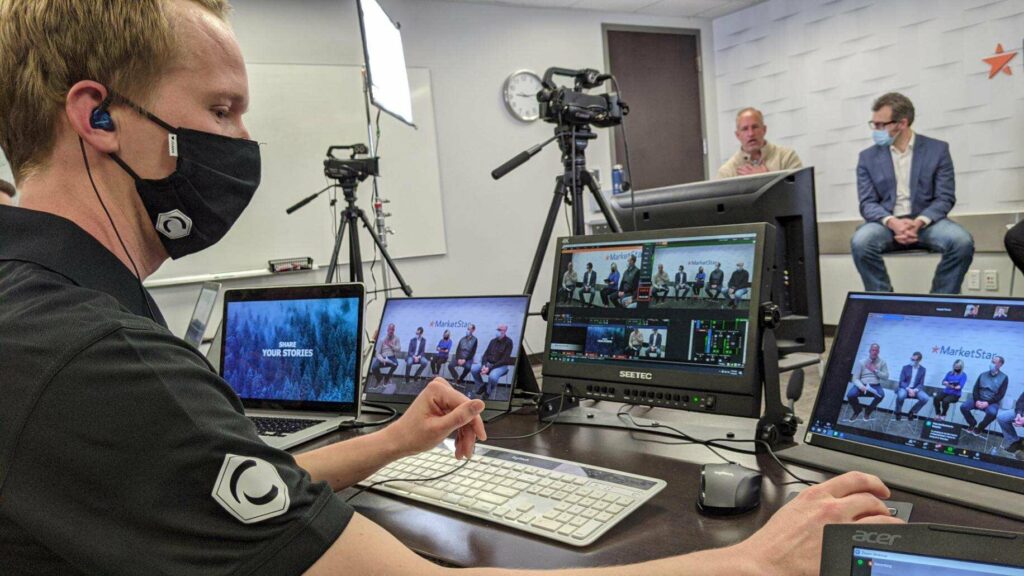 A man in a black mask using several monitors to livestream a panel of people talking.