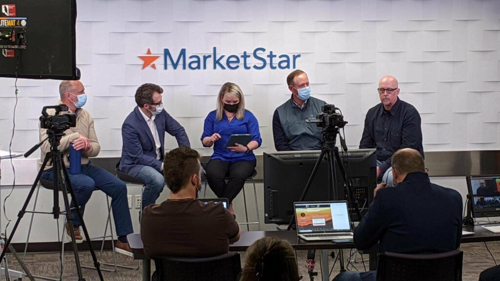 Men behind a table with laptops and a tripod livestreaming a panel of five men and a woman in front of a wall that says MarketStar.