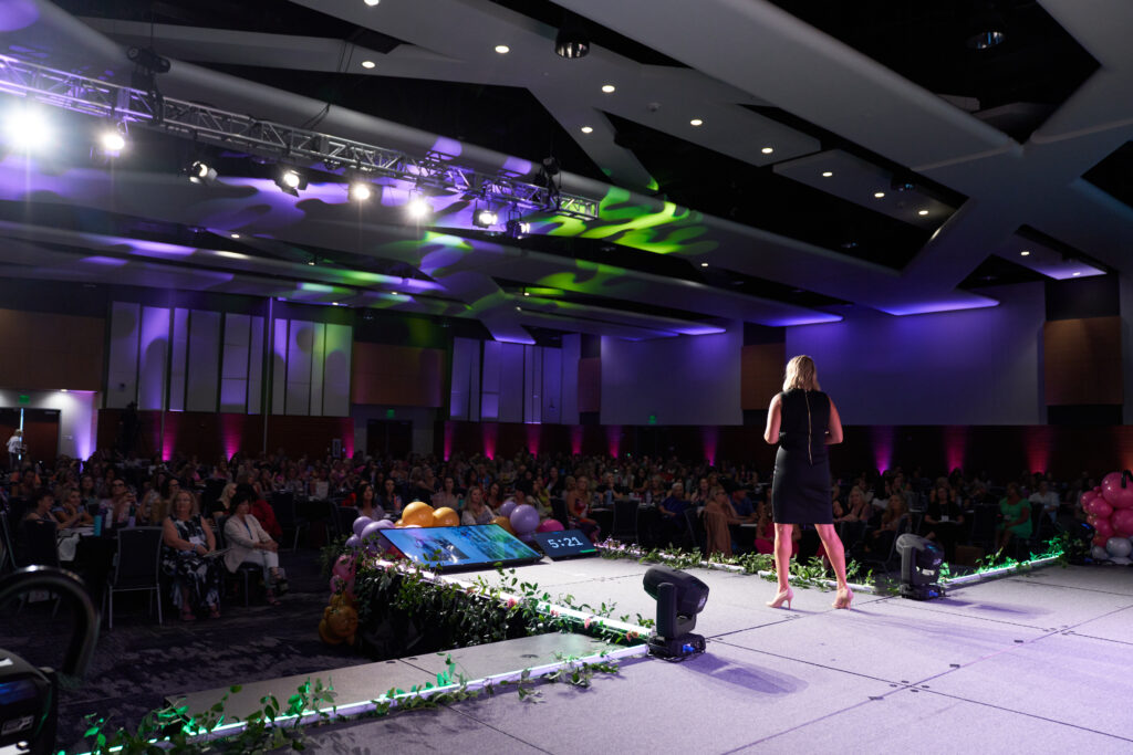 Speaker on large stage in the Grand Ballroom at Utah Valley Convention Center during an event. Full AV purple and green lighting done by Charisma Event Productions.
